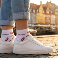 Person iført hvide ankelsokker med røde hjerte-Dannebrogsflag og 'DENMARK' tekst. Sokkerne bæres i hvide sneakers og oprullede jeans på brosten i et solrigt Nyhavn, København