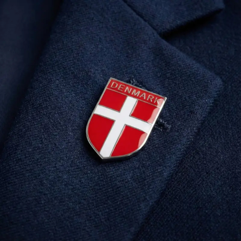 Nærbillede af en blank, skjoldformet metal-pin med det danske flag og teksten 'DENMARK' øverst, fastgjort på reversen af en mørkeblå tekstureret blazer.