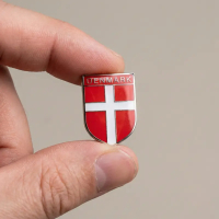 En hånd holder en lille, skjoldformet Dannebrog-pin mellem tommel- og pegefinger mod en neutral baggrund, hvilket demonstrerer dens kompakte størrelse