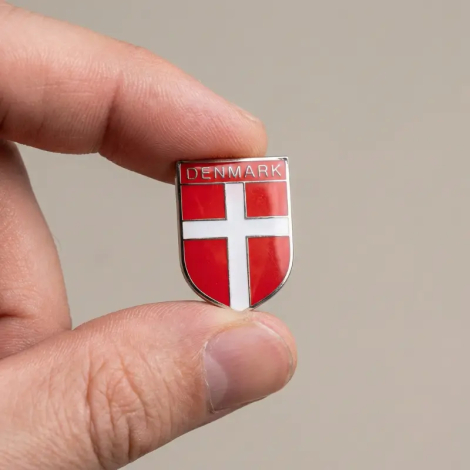 En hånd holder en lille, skjoldformet Dannebrog-pin mellem tommel- og pegefinger mod en neutral baggrund, hvilket demonstrerer dens kompakte størrelse