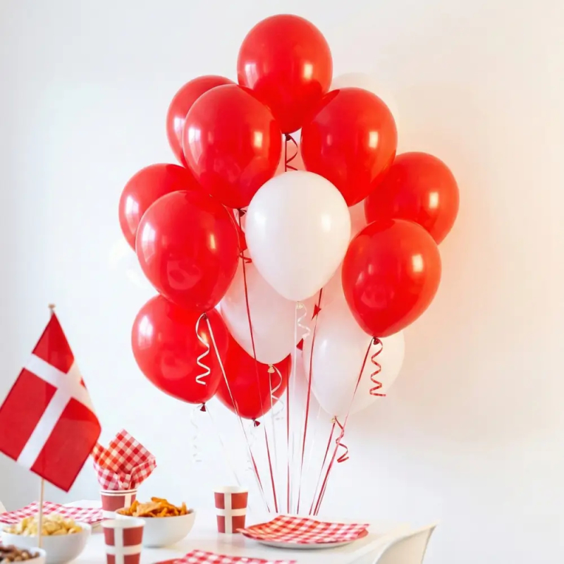 En festlig buket af blandede røde og hvide heliumballoner med krøllet bånd svæver over et bord dækket til dansk fest. På bordet ses et lille Dannebrog-flag, rød- og hvidternede servietter, krus med flagmotiv samt skåle med snacks, alt sammen mod en hvid væg