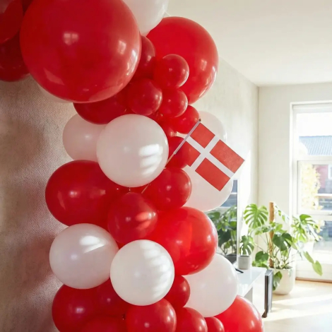 Et udsnit af en organisk ballonbue, sammensat af mange røde og hvide balloner i forskellige størrelser. Et lille dansk flag er stukket ind i ballonguirlanden. I baggrunden ses en lys stue med en stor grøn plante og lysindfald fra et vindue