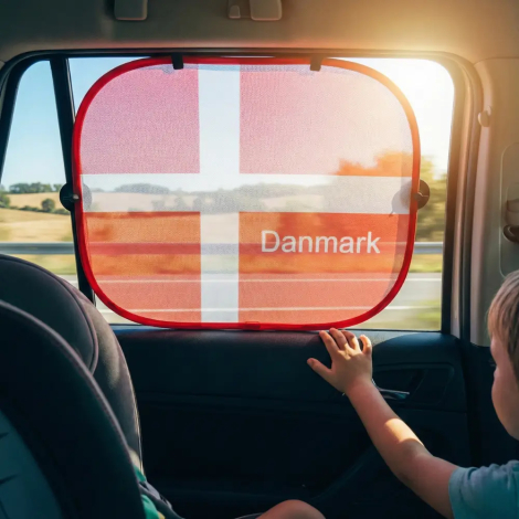 Set indefra bilen: En solskærm med dansk flag monteret på ruden ved siden af et barn i autostol. Billedet viser, hvordan mesh-nettet skygger for solen, men stadig tillader udsyn til landskabet.