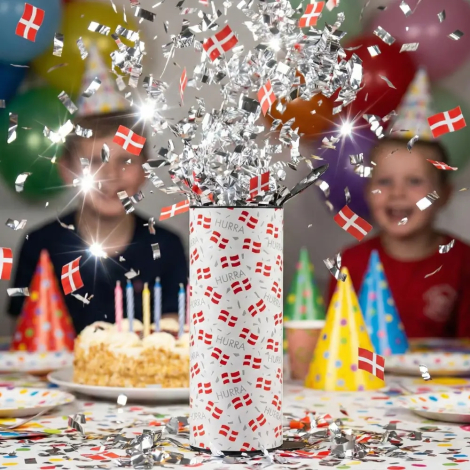 Eksploderende HURRA bordbombe i festligt miljø. En sky af skinnende sølvkonfetti og små danske papirflag skydes op i luften over et fødselsdagsbord
