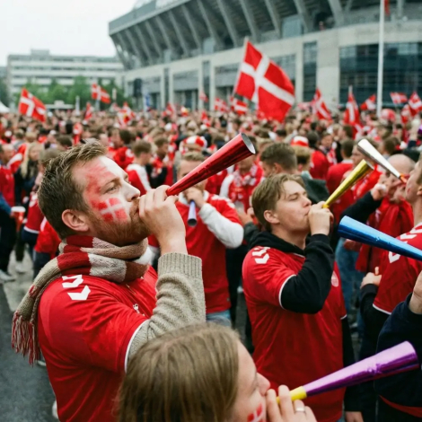 En stor forsamling af danske roligans foran Parken. I forgrunden står en mand i profil og puster i et langt rødt fest-horn, mens andre fans omkring ham puster i blå og gule horn. Stemningen er præget af fest og kaos før kampstart.