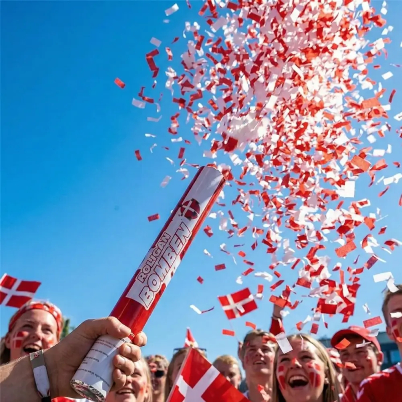 En hånd holder et 'Roligan Bomben' konfettirør op mod en klar blå himmel, idet det affyres og sender en massiv sky af rød og hvid papirkonfetti op i luften.
