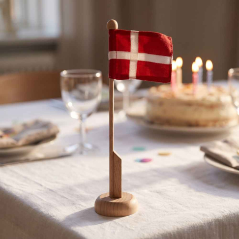 Stemningsbillede af det 25 cm høje Dannebrog bordflag i træ på et festligt dækket fødselsdagsbord med hvid dug, vinglas og en fødselsdagskage med levende lys i baggrunden.
