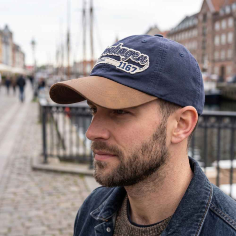 Nærbillede af en mand iført en navy kasket med brun skygge og broderet 'Copenhagen 1167'-logo, stående i et afslappet bymiljø med en sløret havn i baggrunden